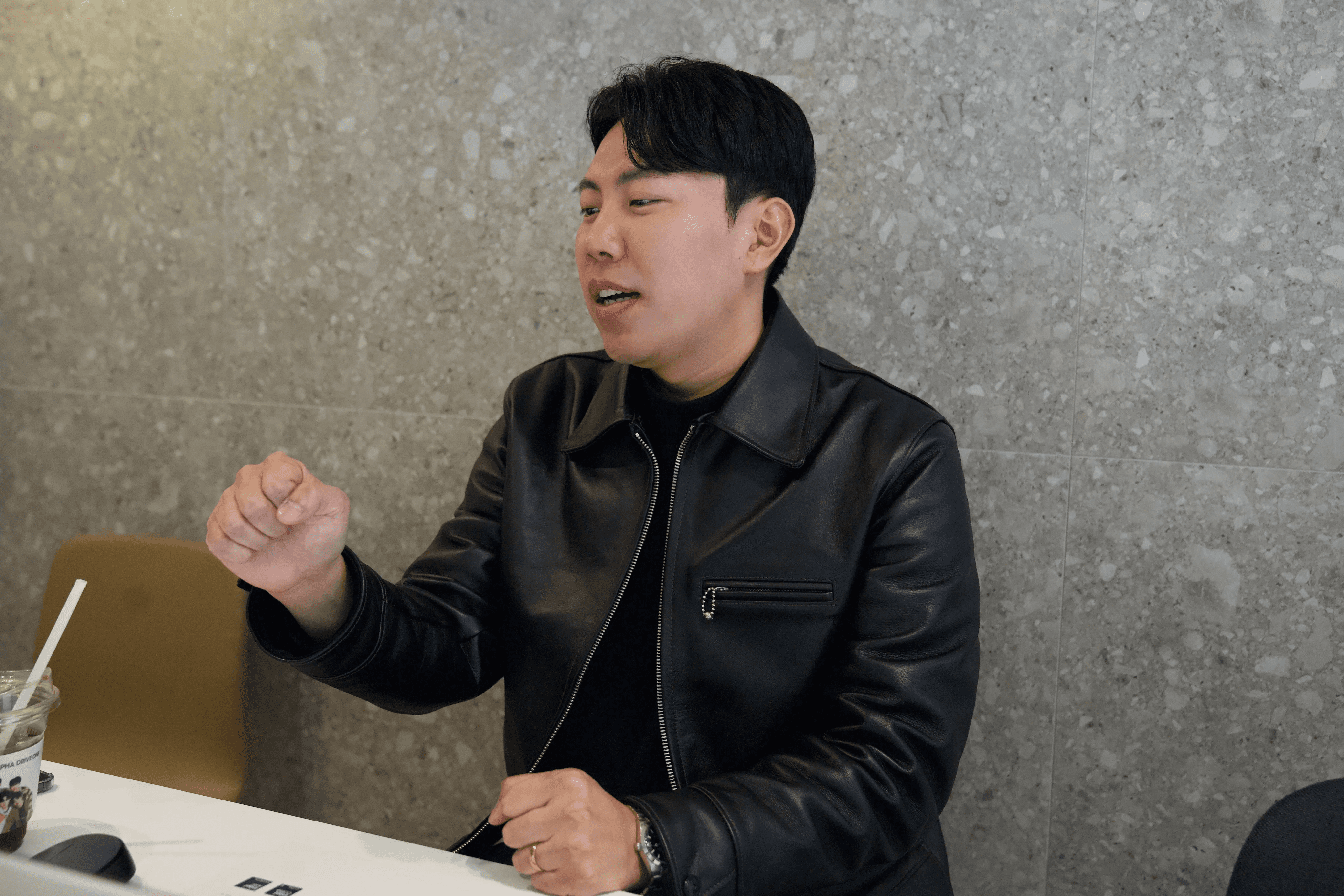 회색 테라조 벽을 배경으로 검정 가죽 재킷을 입은 남성이 오른손 주먹을 쥐며 강조하는 제스처를 취하고 있음. 테이블 위에 음료컵이 놓여 있으며 발언 중인 표정.