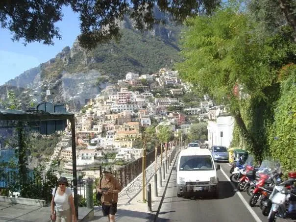 The narrow Amalfi Coast road with a car, parked scooters and people walking.