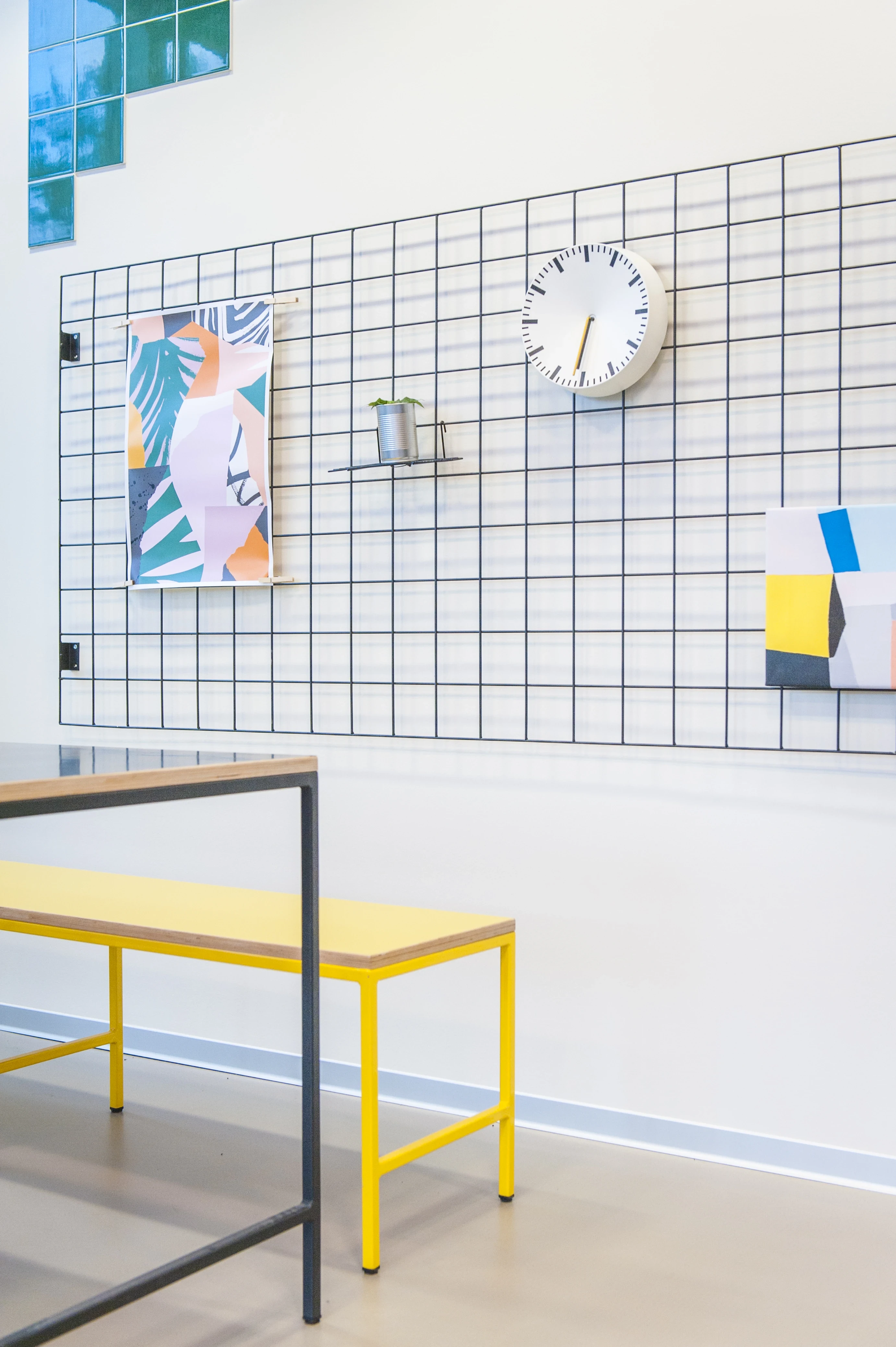 Wire grid wall panel at KANTINE KOMTUR with a round white clock, a tropical print and a small shelf, next to a yellow bench and green pixel tiles.