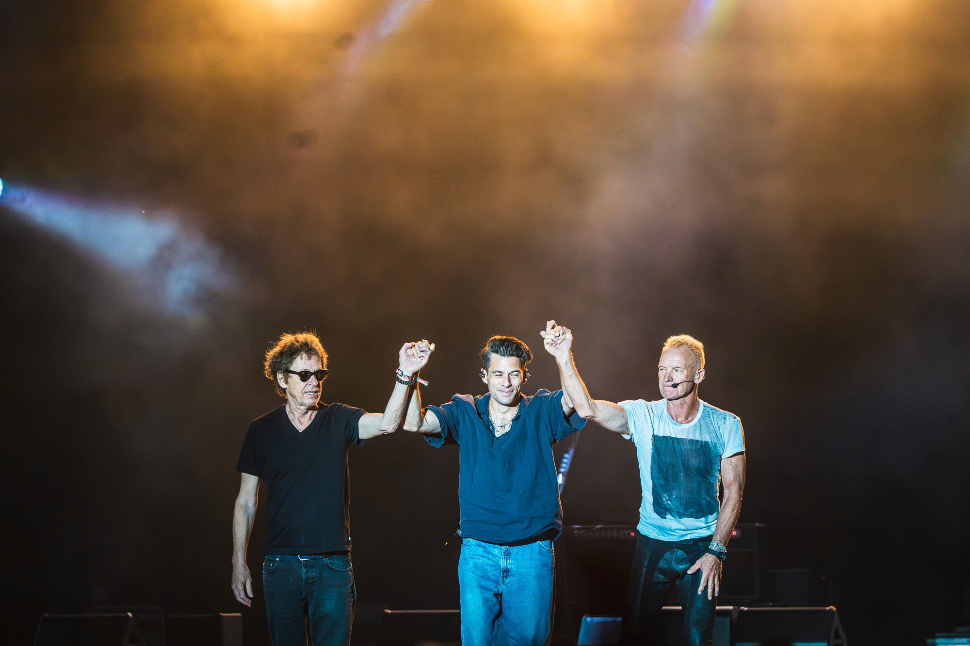 Trois musiciens se tenant la main, bras levés, salut final sur scène à contre-jour