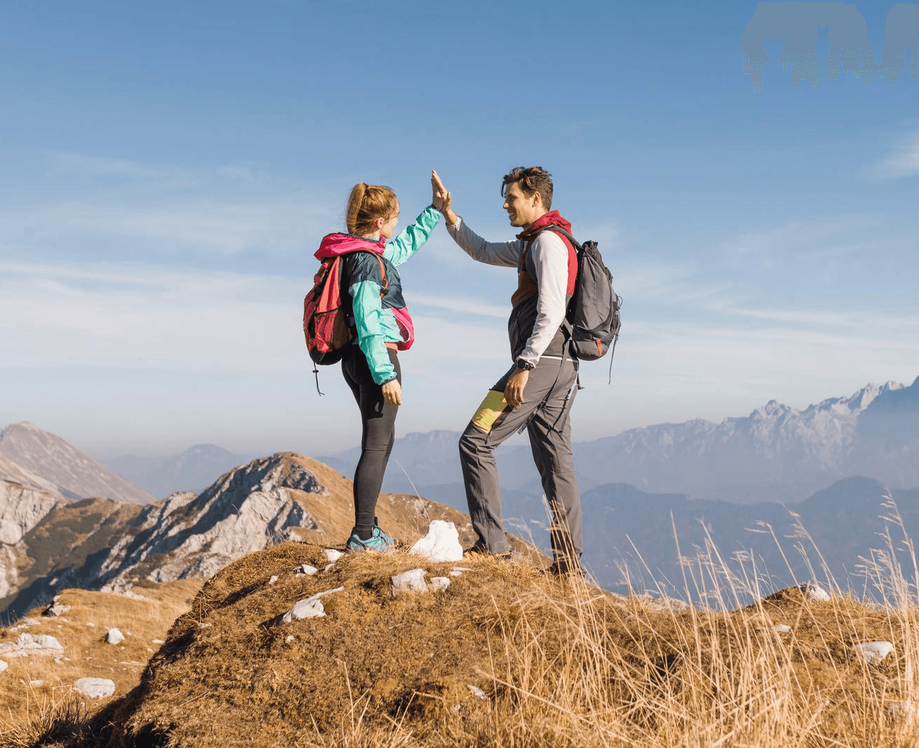 A man in an orange raincoat helping a woman in a yellow jacket step across rocks at a mountain lake.