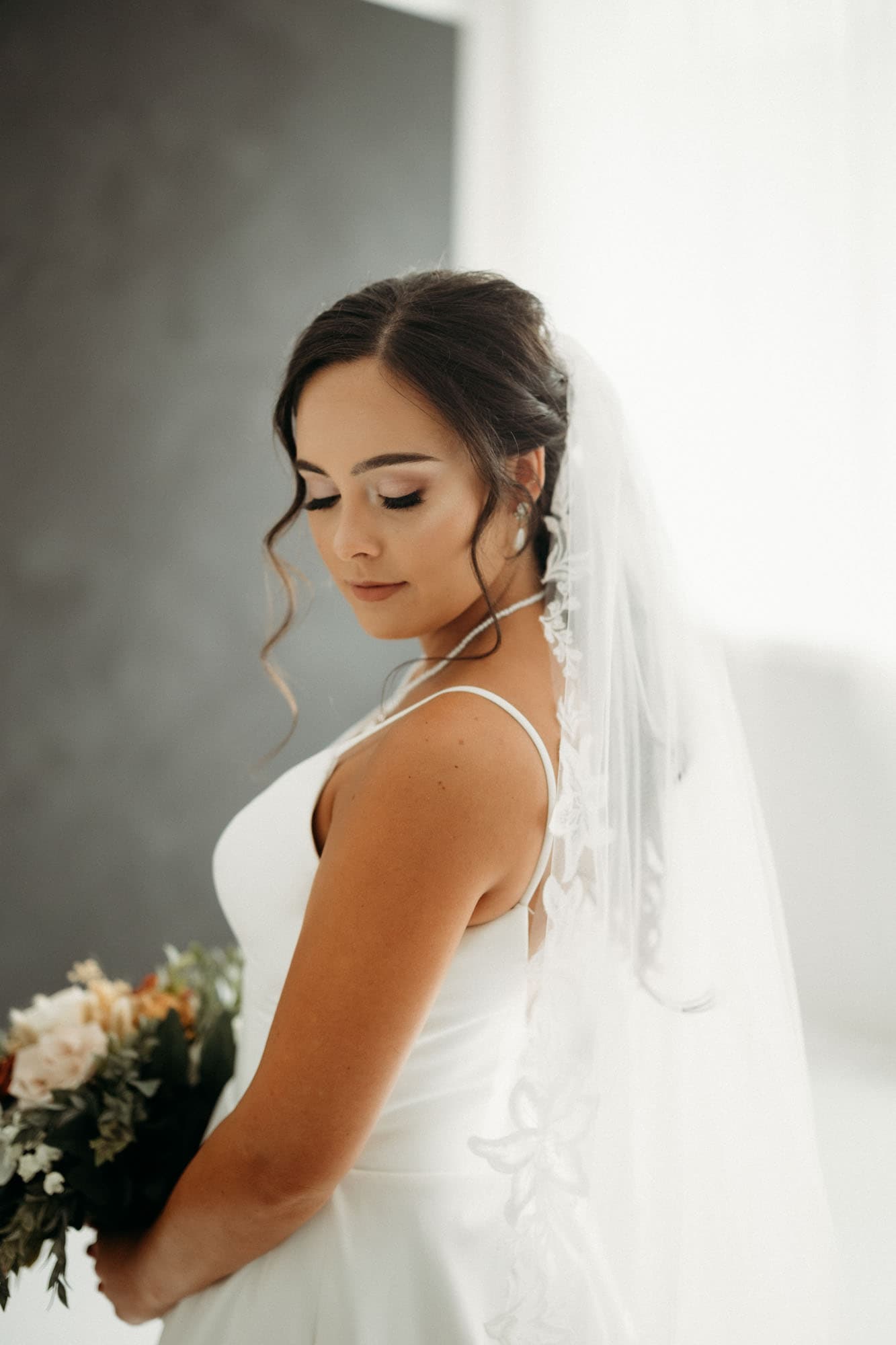 The bride stands against a dark backdrop, holding her bouquet and gazing down, captured at Revelator Studio, a natural light studio in Shreveport.