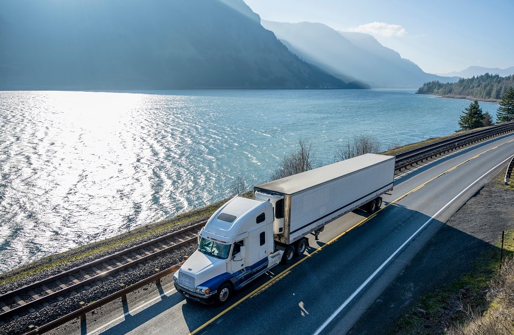 Aerial fisheye-lens photo with a blue-toned filter showing a five-axle refrigerated truck on a coastal road. The truck has a white cab with a blue stripe near the wheels and an aerodynamic accessory on the roof. A railway line runs between the road and a body of water bordered by mountain slopes. The image represents BeyondTrucks' Dry Van and Reefer Solution for large private fleets and for-hire carriers.