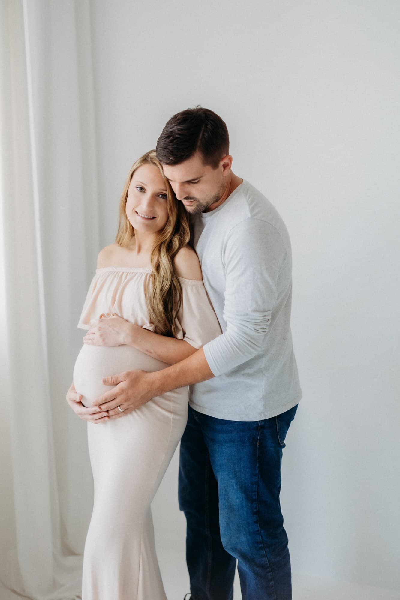 A couple embraces in a soft, natural light setting at Revelator Studio in Shreveport, capturing a serene maternity moment.