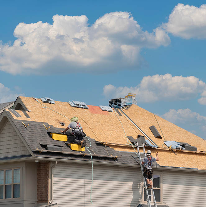 Workers install roofing materials on a house under a blue sky with fluffy clouds.