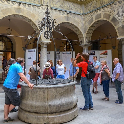 Туристическа група се събира около изящен каменен кладенец в двора на историческа сграда със сводести стени и плакати на заден план.