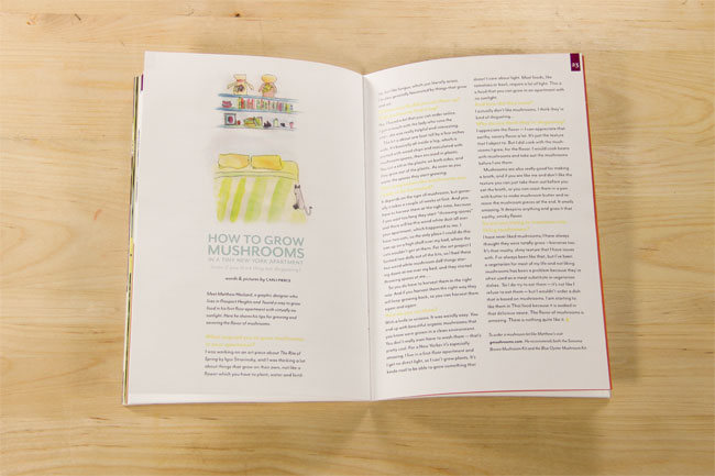 The image shows an open book resting on a light wooden surface. The left page displays a colorful illustration and text titled "HOW TO GROW MUSHROOMS IN A TINY NEW YORK APARTMENT," accompanied by a small graphic of a cat peeking out from behind a mushroom. The right page contains text divided into paragraphs. The page numbers visible are 55 and 56. The book's spine is red, and the pages have a cream or off-white background.