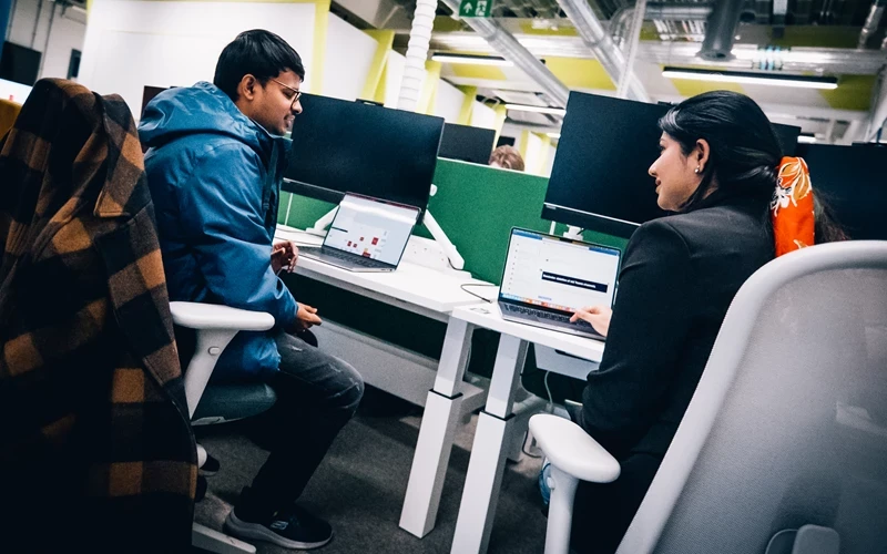 Two people work at adjacent desks in an open‑plan office, seated in ergonomic chairs and using laptops with privacy screens. One person wears a blue jacket and leans forward in conversation, while the other types at their desk. Divider panels, exposed ceiling pipes, and overhead lighting create a modern workplace environment focused on collaboration and individual work.