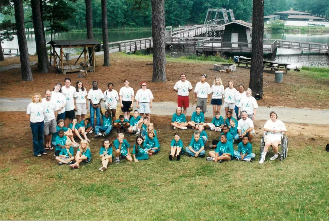 Large group photo of campers and staff. They are wearing teal and white Camp Juliena shirts and sitting/standing on the grass.