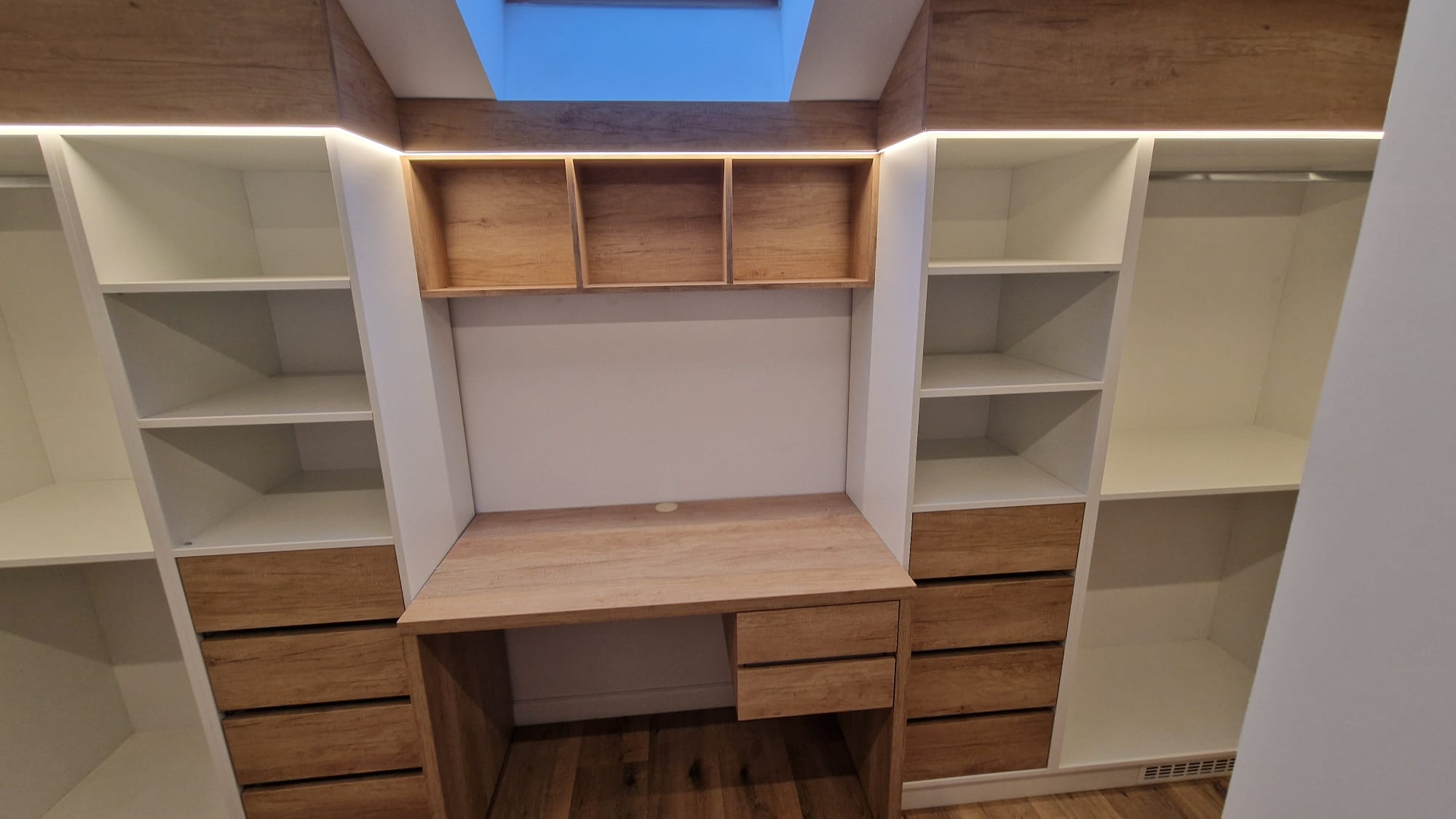 Walk‑in closet simetric cu insulă de birou centrală, module albe și sertare din lemn sub fereastra Velux.