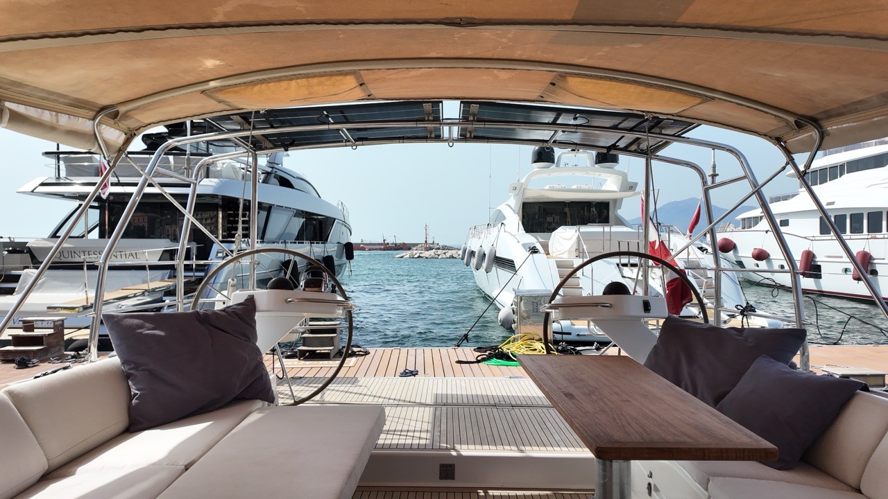 View across the Hanse 575's cockpit beneath its tan bimini, showing twin wheel stations, white cushioned seating, a teak table, and a marina full of motor yachts beyond the swim platform.