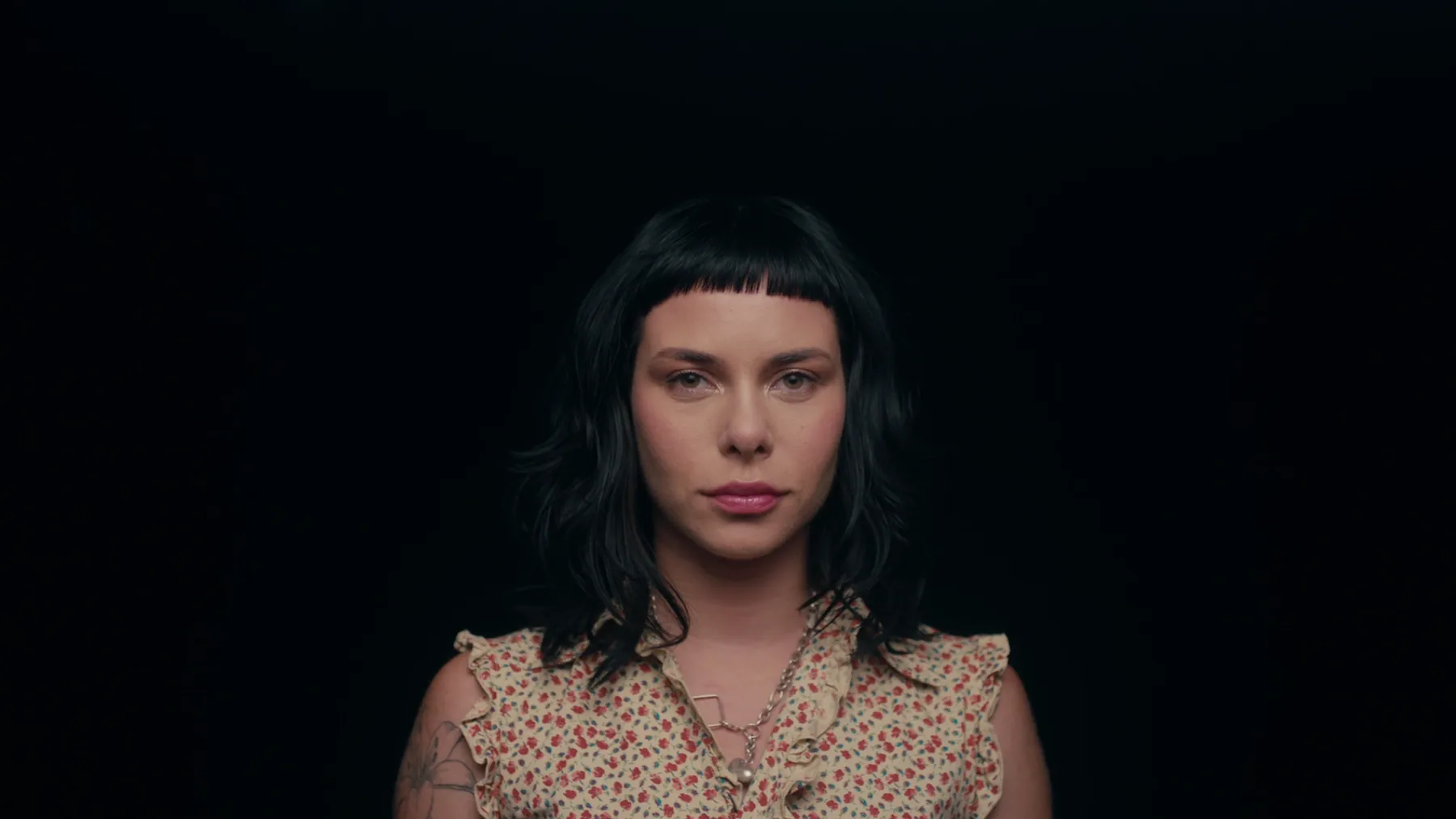 Portrait of a woman with dark hair and blunt bangs in a cream floral top, lit with neutral warm skin tones against a pure black background.