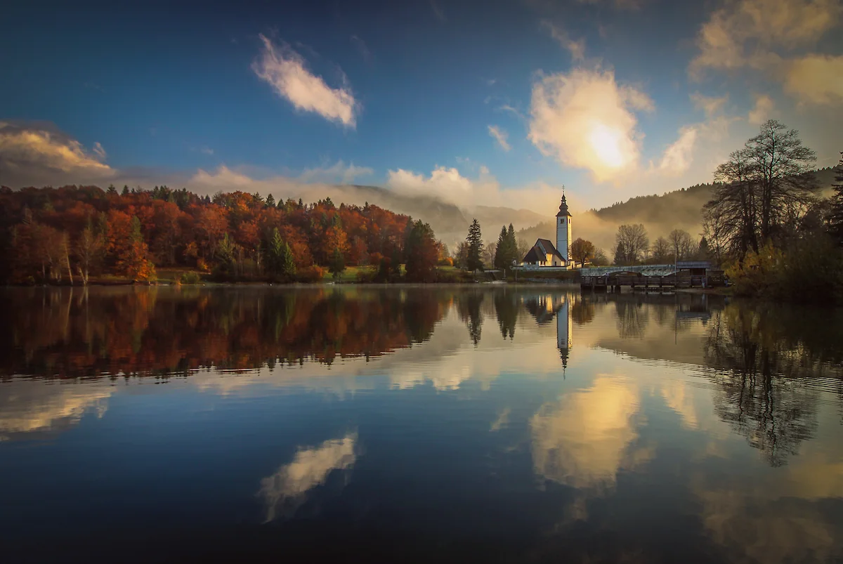 A church surrounded by autumn colored forest reflecting in Lake Bohinj, Slovenia, on a foggy blue sky morning.