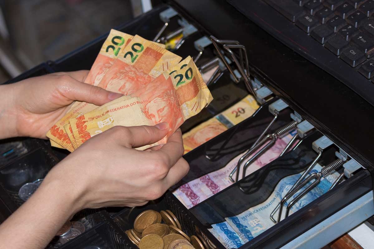 Mãos segurando cédulas de real sobre gaveta de caixa com notas e moedas organizadas.
