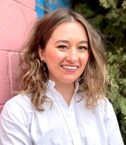 A smiling person with curly hair wearing a white shirt, standing against a colorful wall with greenery.