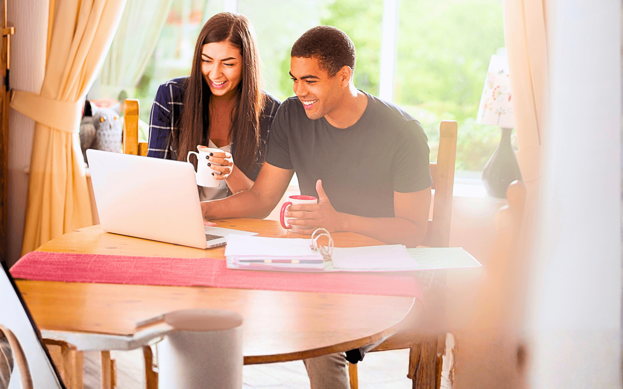 Couple smiling while looking at a laptop computer together at a round wooden table, holding mugs and documents.
