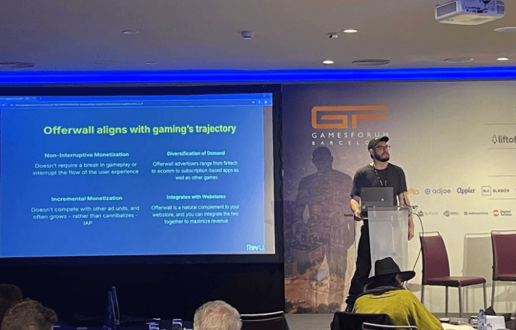 Speaker giving a keynote presentation on a stage next to a projector screen titled "Offerwall aligns with gaming's trajectory."