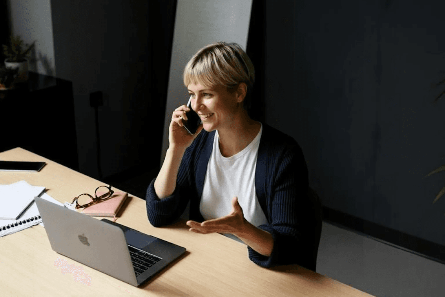 Woman with blonde hair smiling while talking on the phone at a desk with a laptop and papers.