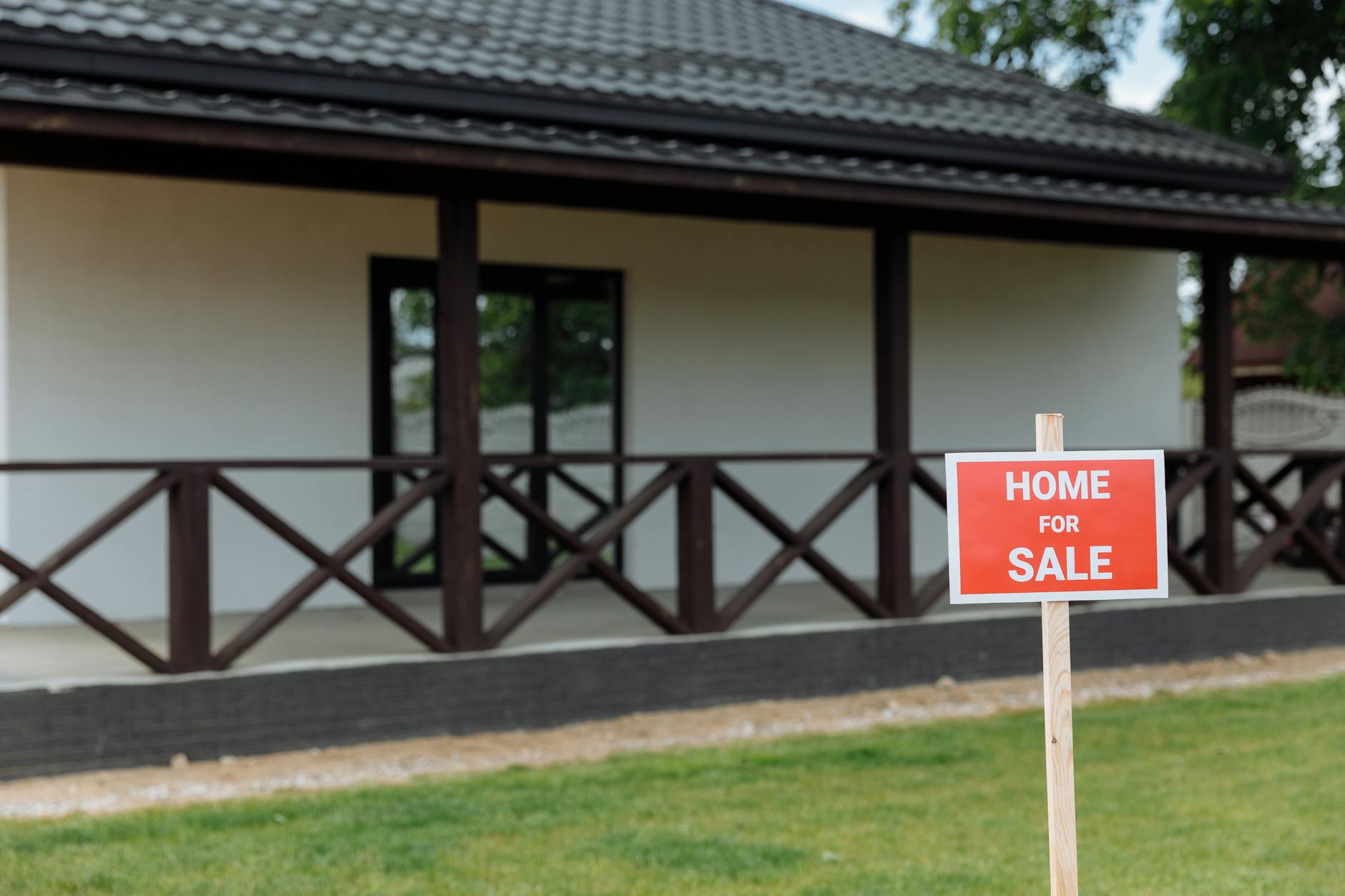 Red Home For Sale sign on a lawn in front of a white bungalow with brown cross-hatch wooden fence detailing