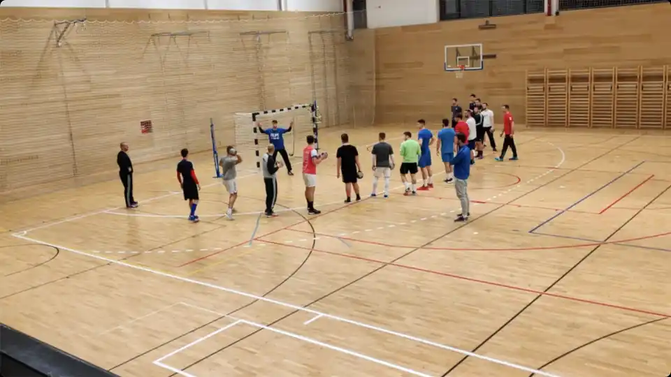 Handball players holding balls during a group shooting drill, with a coach beside the goal and a goalkeeper ready to face the shots.