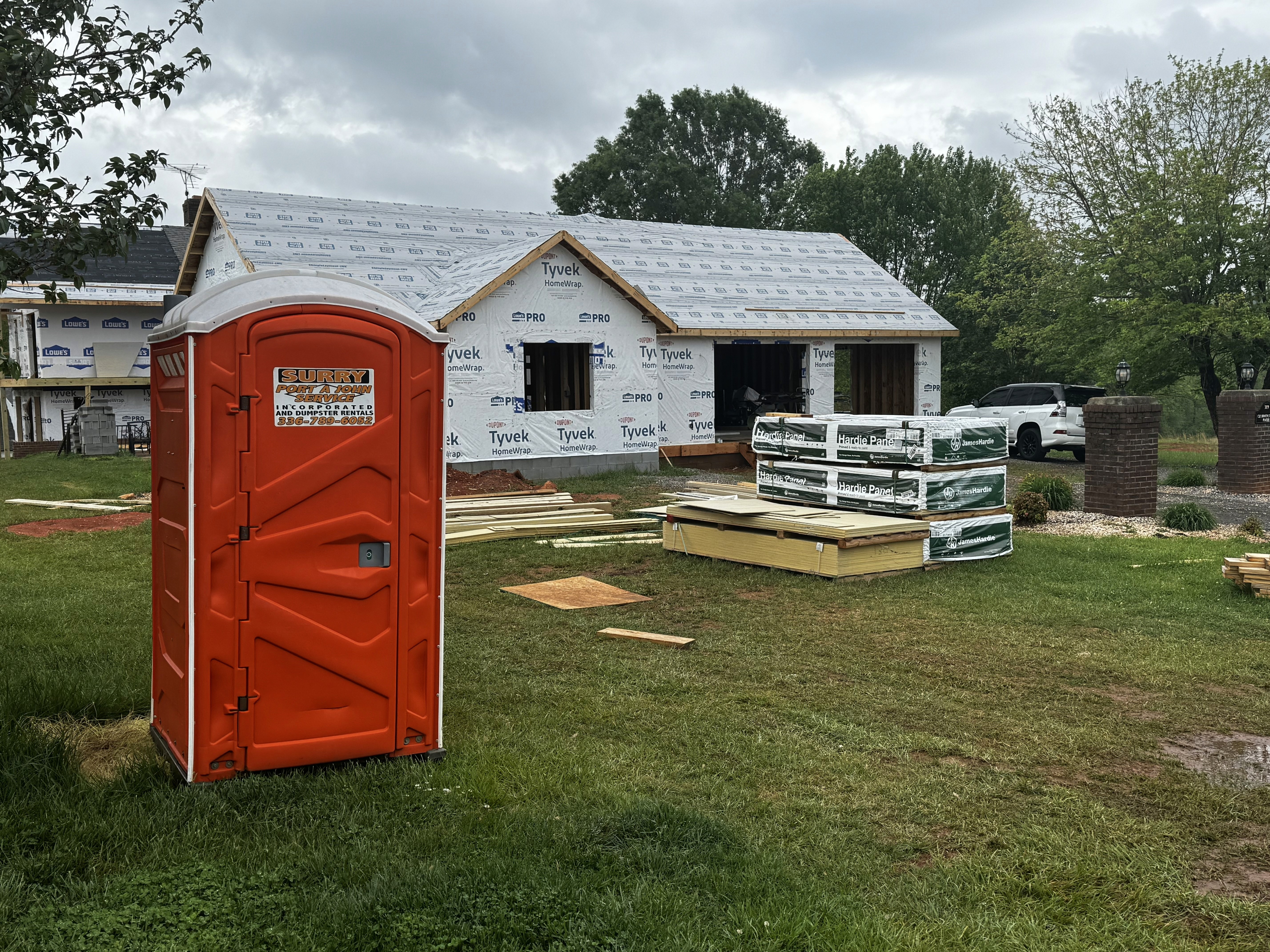 A partially constructed house wrapped in Tyvek is surrounded by stacks of building materials and lumber on a grassy lawn, with an orange portable toilet in the foreground and a parked car in the background.