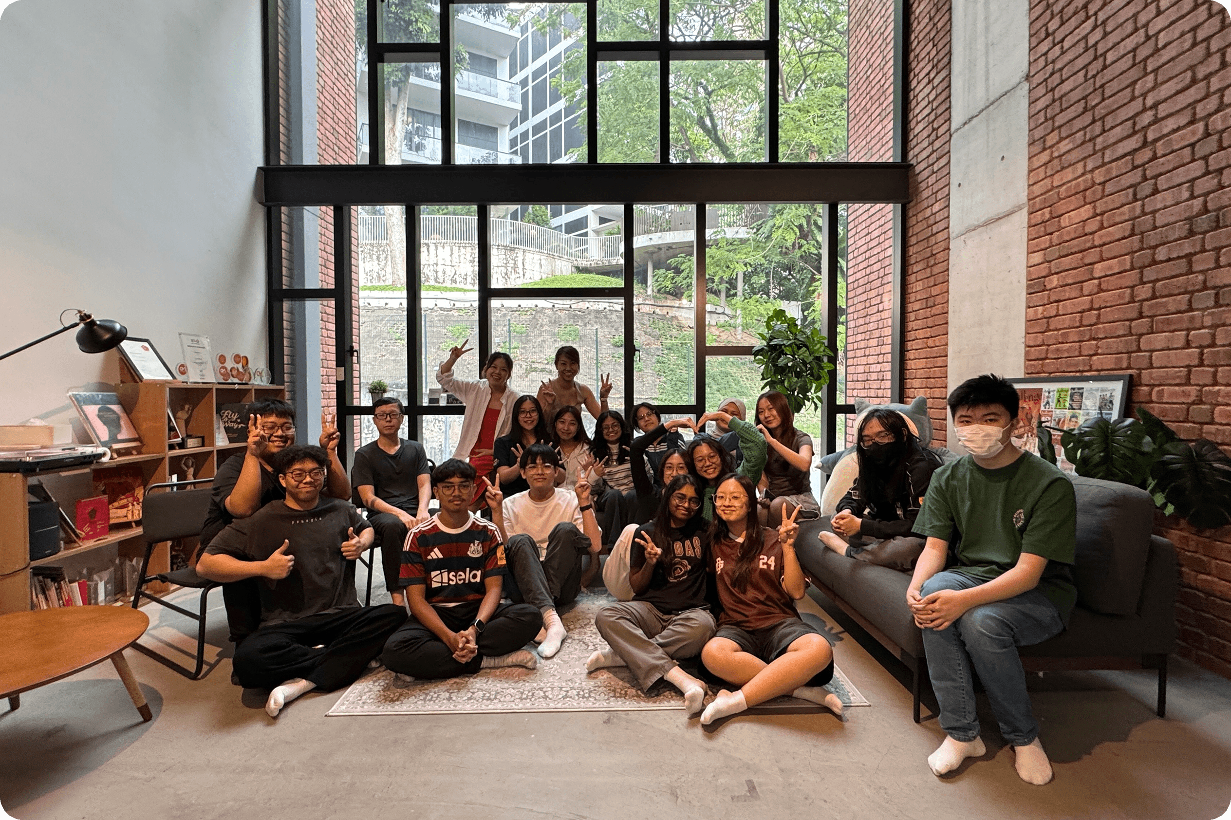 A group photo of 19 students sitting in front of floor-to-ceiling glass windows