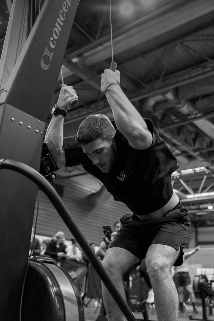 A shirtless man intensely using battle ropes in a gym, with another man in the background.