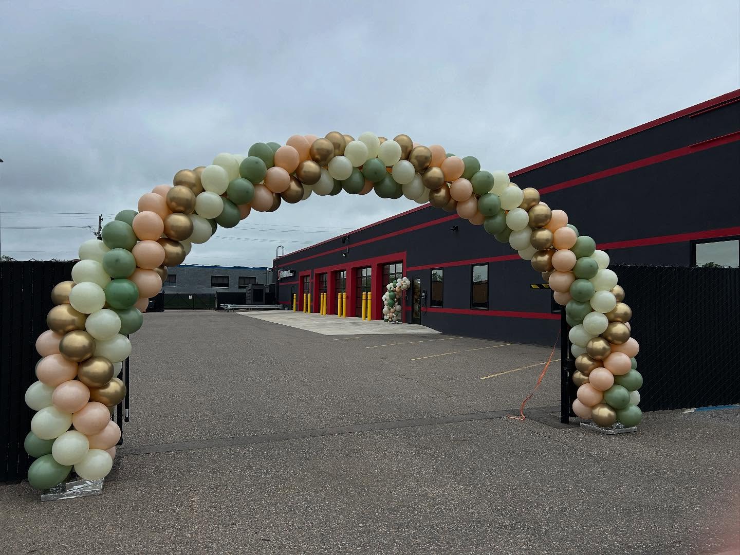 An arch of colorful balloons, featuring green, gold, and peach hues, frames the entrance to an industrial building with dark walls and red accents against a cloudy sky.