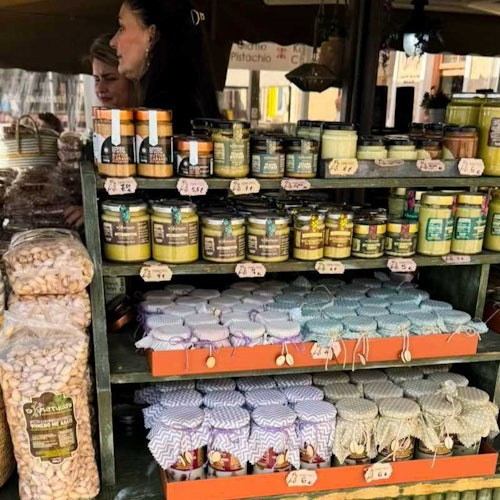 一个市场摊位，货架上陈列着蜂蜜罐、果酱和包装好的坚果。背景中可以看到两个人。