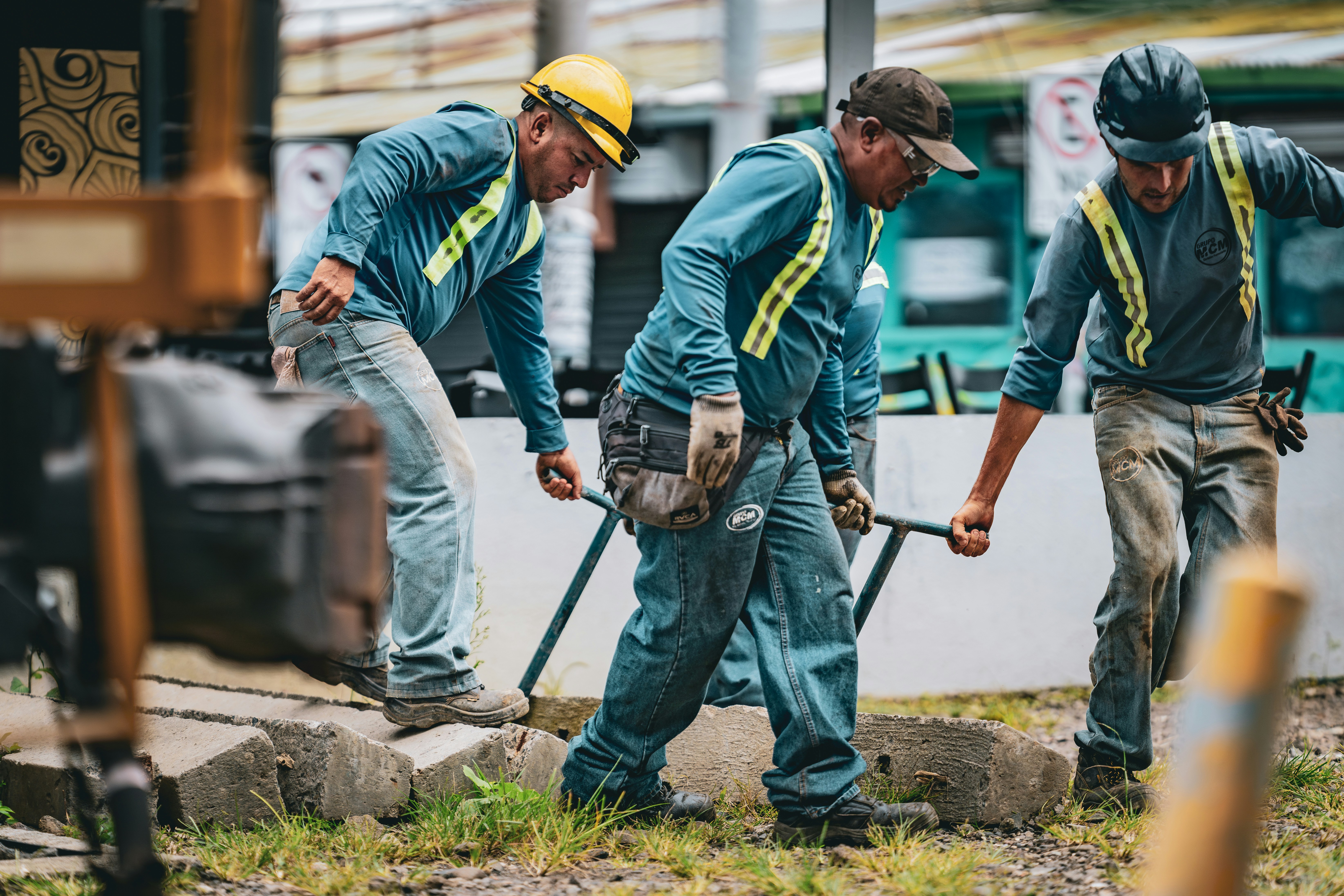 Three trades workers in matching blue work shirts with hi-vis reflective stripes working together to move heavy equipment at an outdoor job site.