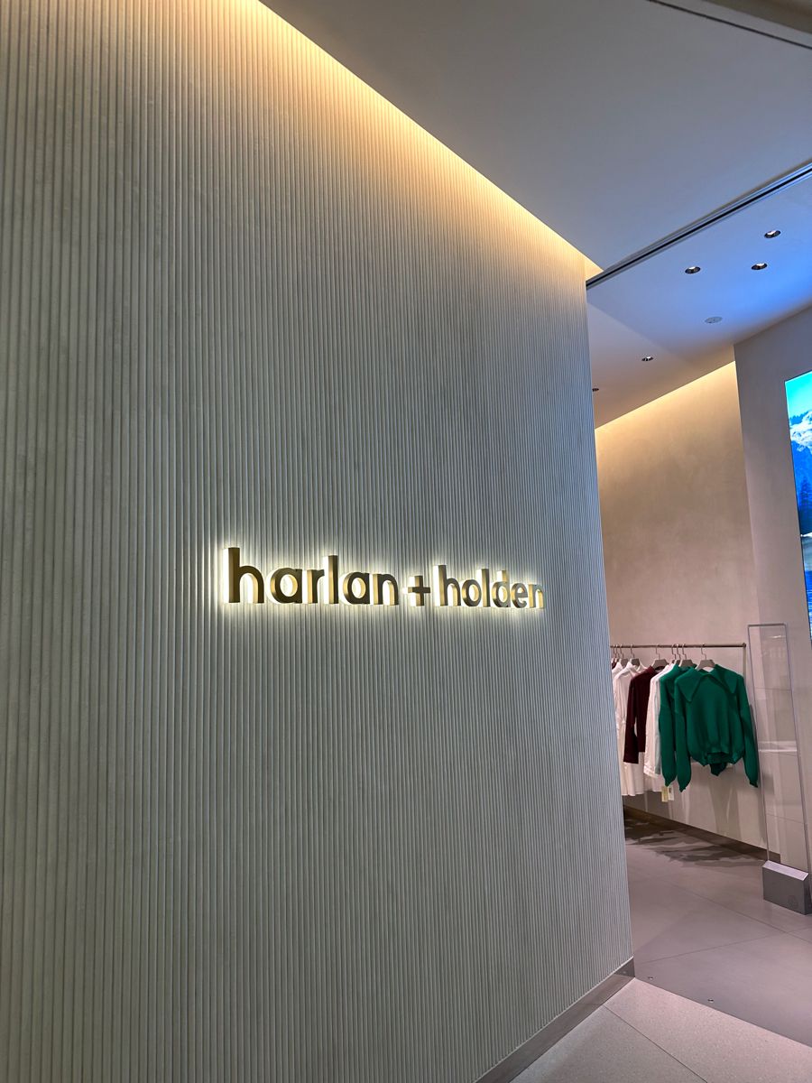 Illuminated “harlan + holden” wall sign inside a minimalist retail store with textured wall panels and clothing displays.