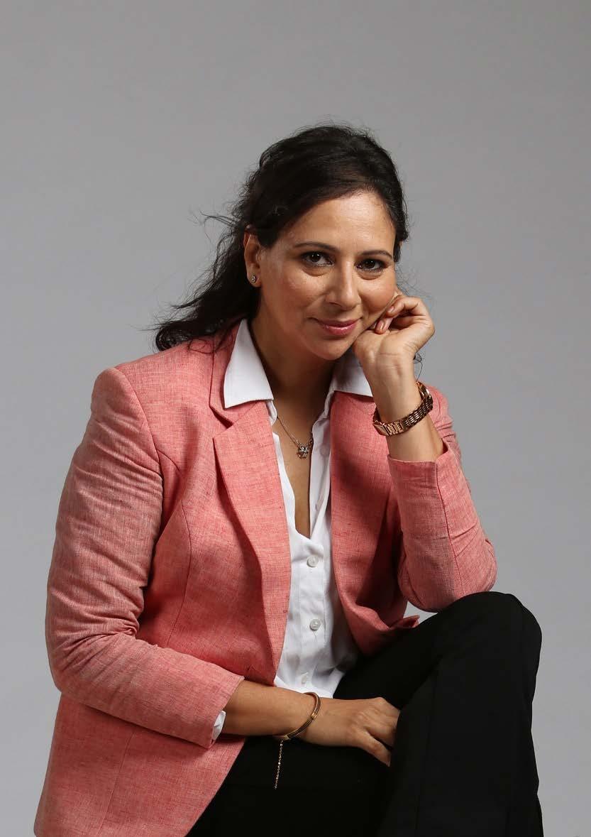 Smiling female therapist with short gray hair, wearing a brown top and a purple shawl, surrounded by a cozy background.