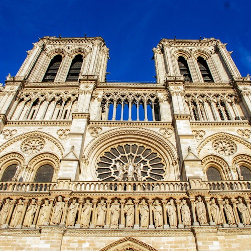 The facade of Notre-Dame Cathedral with twin towers, ornate carvings, a large circular stained glass window, and statues against a blue sky.