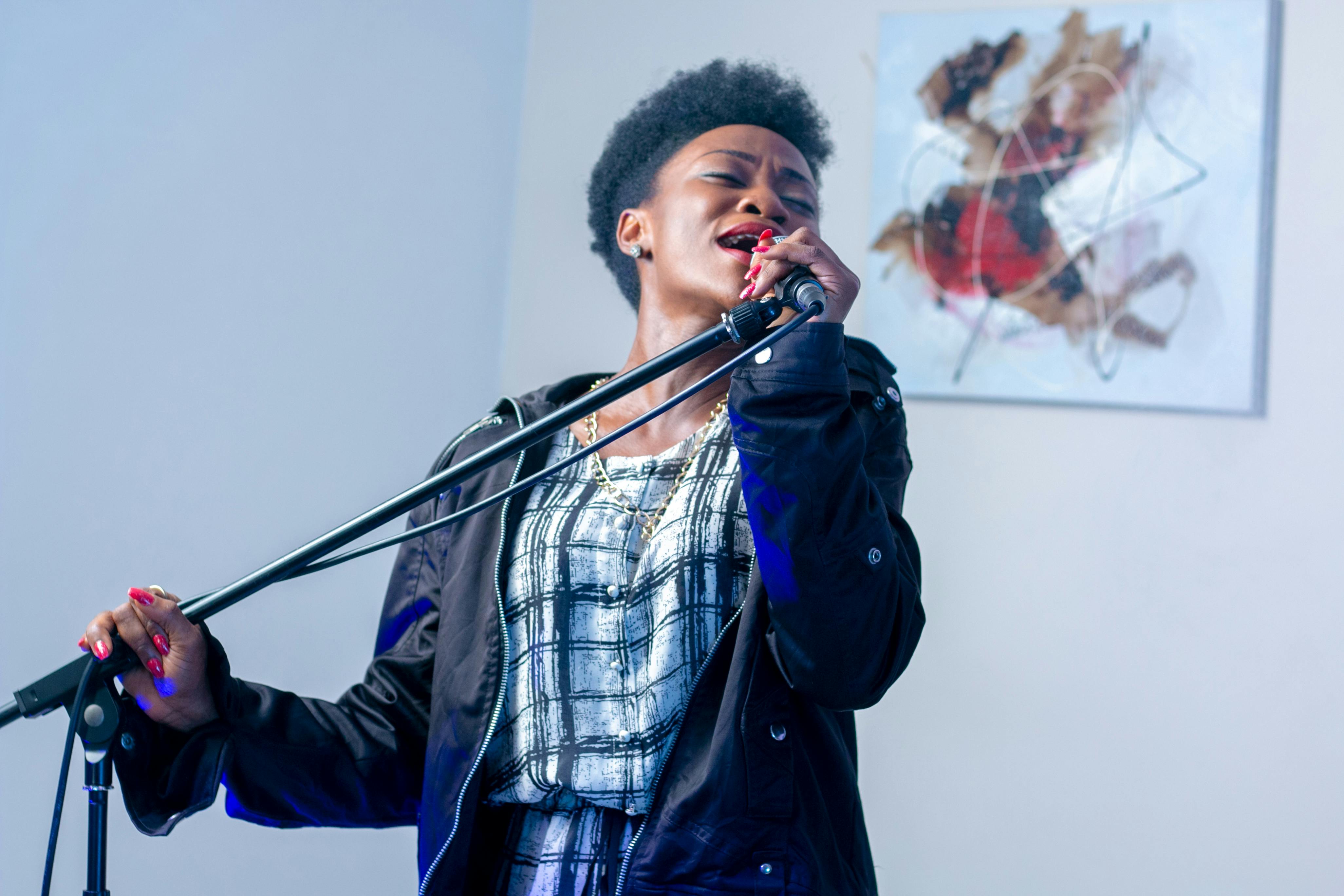 A singer passionately performing into a microphone while holding a stand, wearing a black jacket and patterned outfit, with expressive emotion on her face.