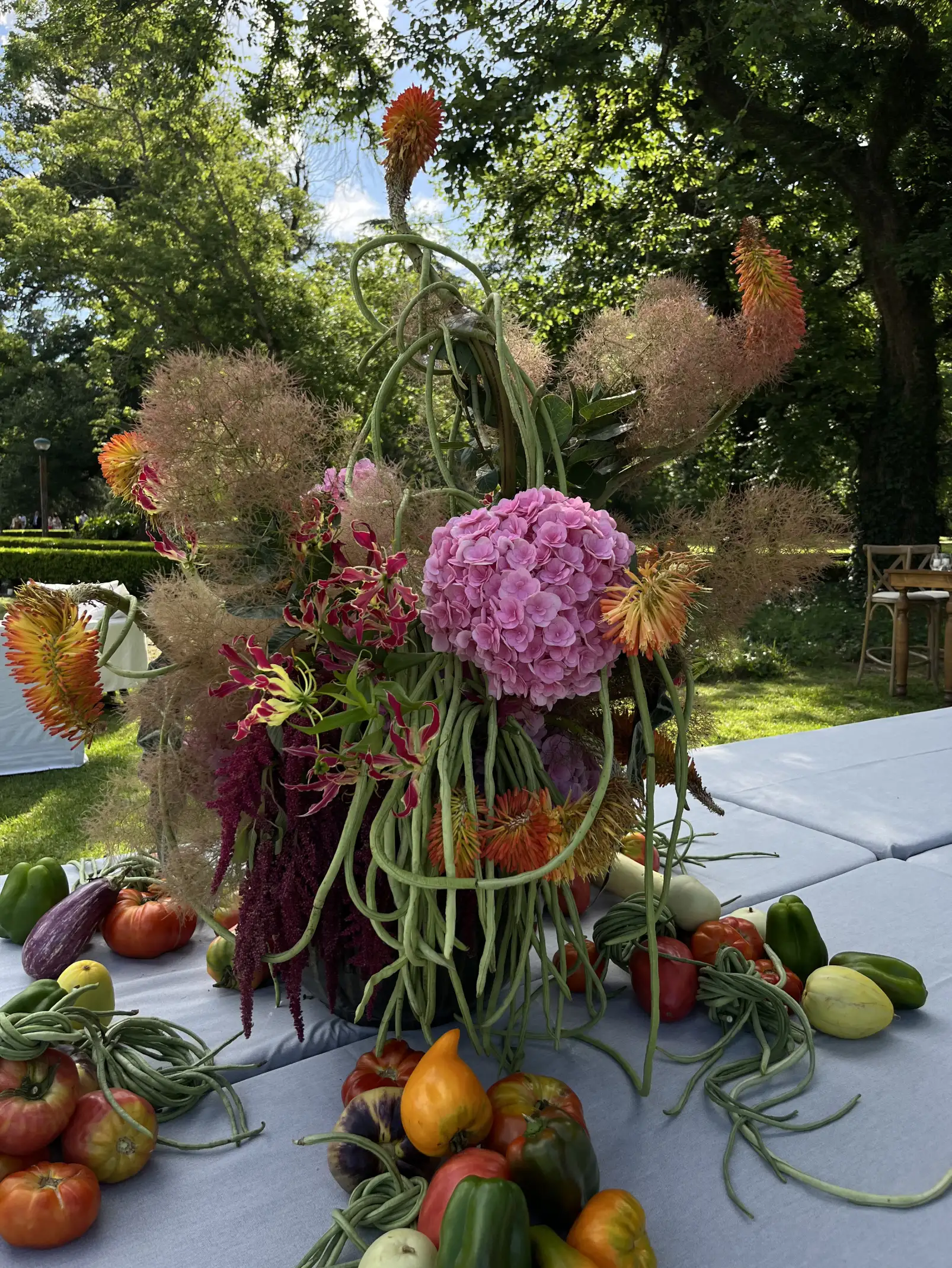 Arreglo floral exuberante con hortensias, gloriosas y follaje, rodeado de vegetales frescos sobre mesa al aire libre.