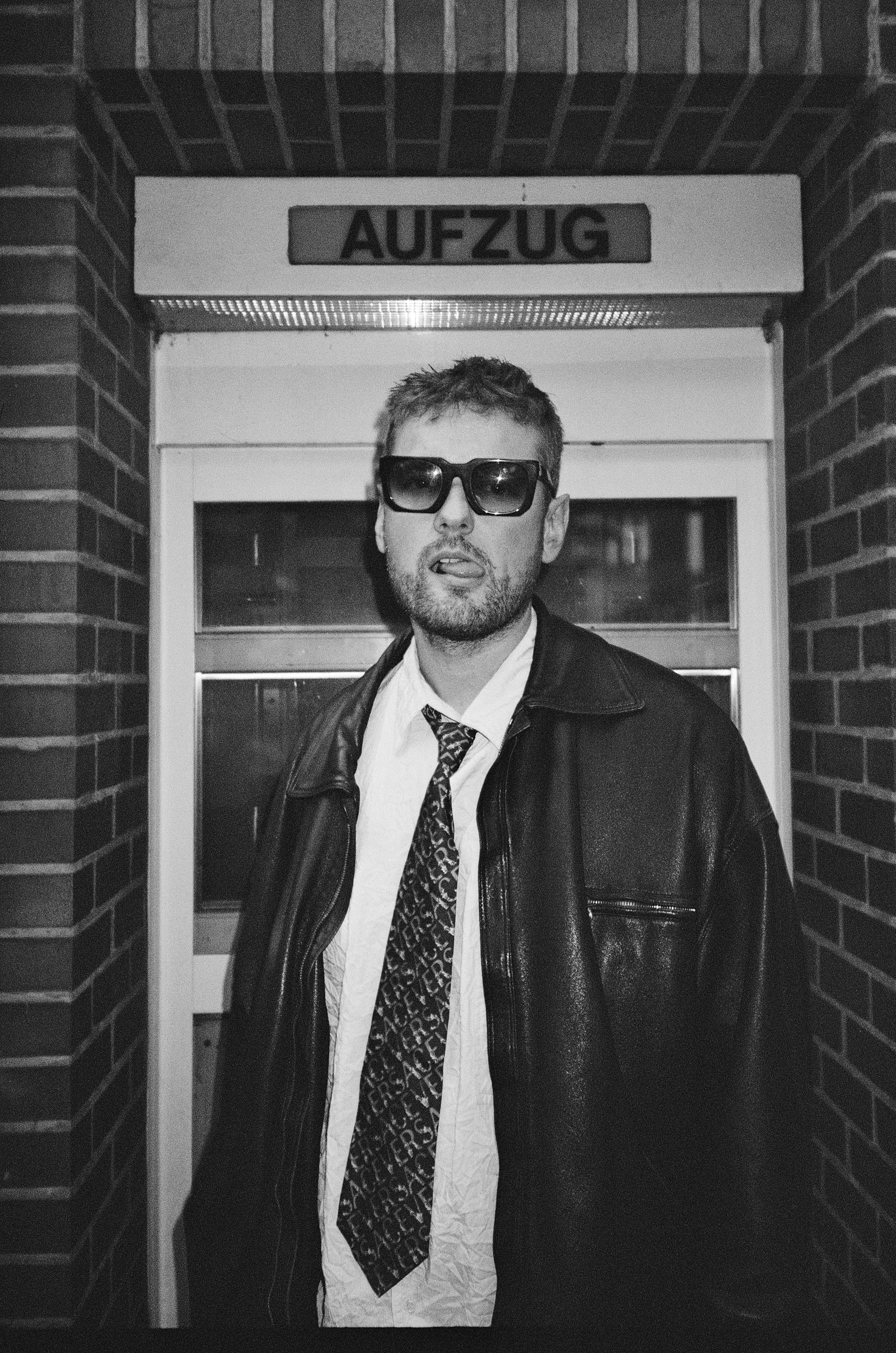 A raw, black-and-white portrait of Mahoni Zingelmann with sunglasses and an undone tie standing provocatively in front of a German elevator sign.
