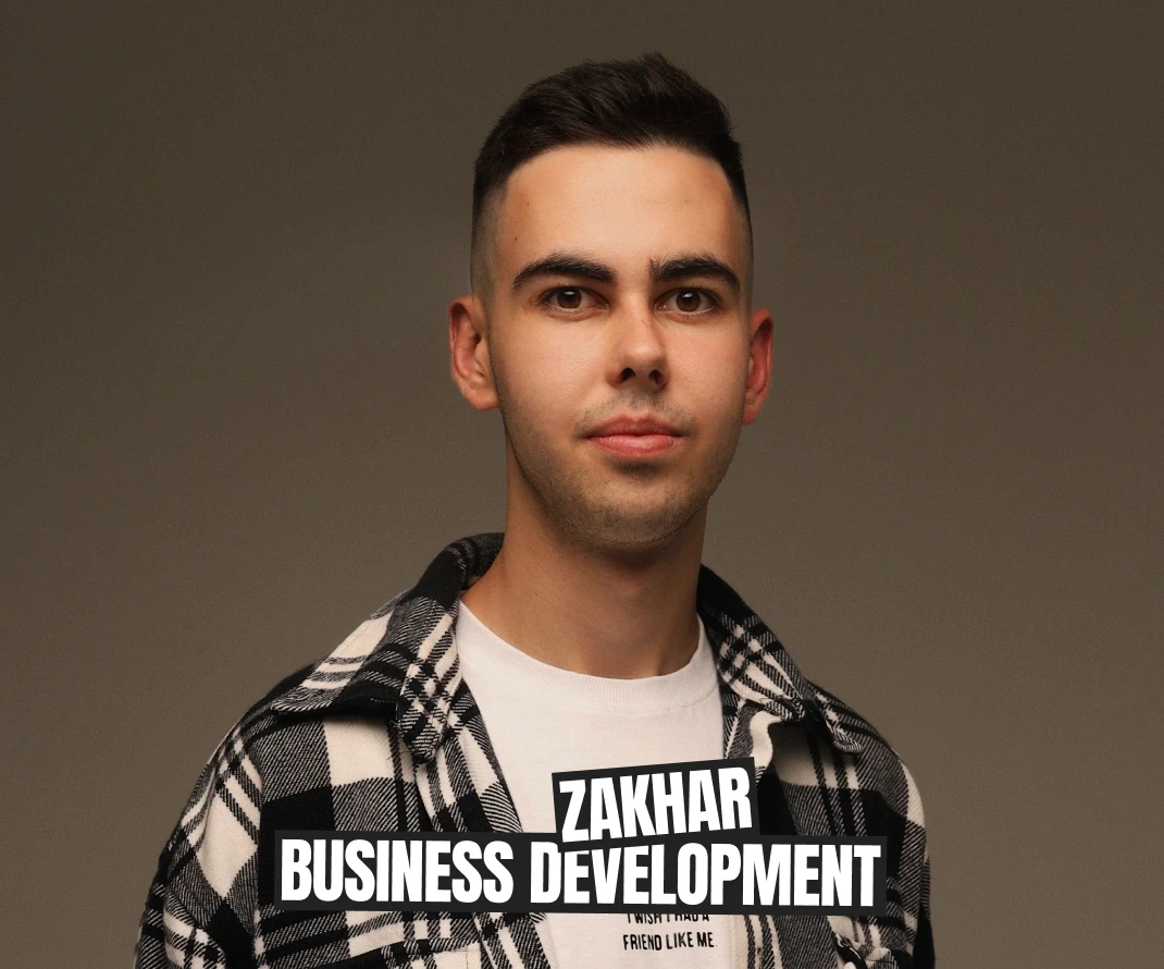  This image features a young man with short, dark hair, dressed in a black-and-white checkered shirt over a white t-shirt. He has a calm and confident expression, and he stands against a plain background, which enhances the focus on him. A text overlay at the bottom of the image reads "ZAKHAR BUSINESS DEVELOPMENT," indicating that his role is related to business development.