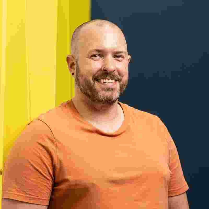 A man in an orange t-shirt with short hair and a short beard smiles at the camera against a yellow and blue background