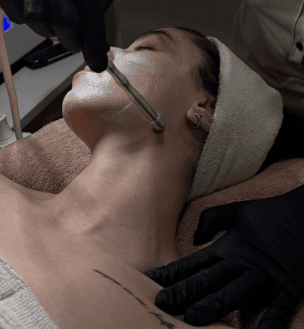 A cozy and inviting spa room with a neatly made bed, soft lighting, and warm wooden tones. The scene is partially framed by green plants, adding a natural and soothing touch.