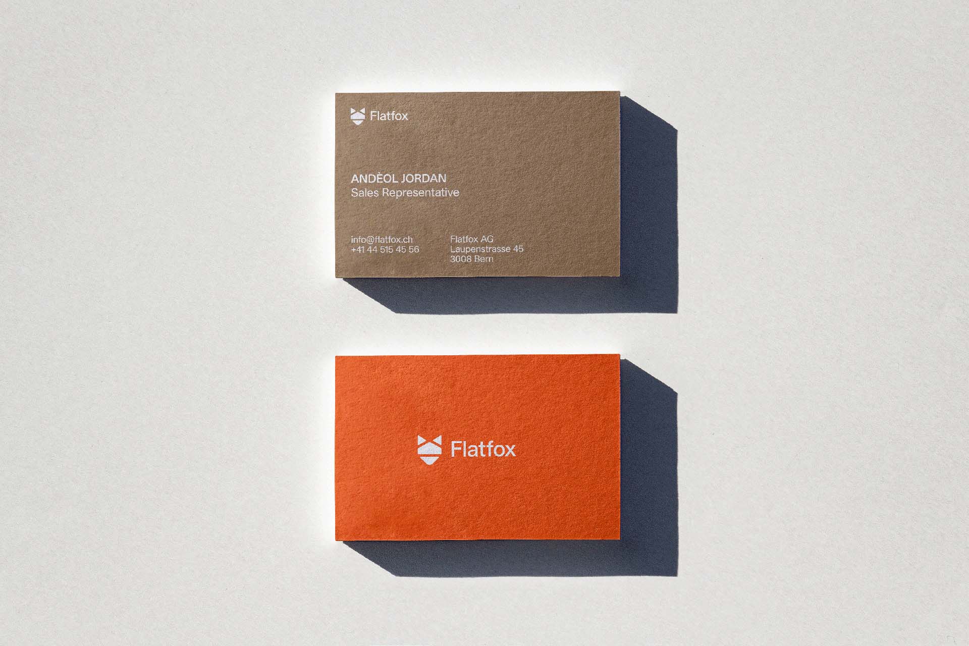 Two professional business cards for a Sales Representative at Flatfox, one in earth-toned grey and the other in vibrant orange. The stationery design uses structured Swiss typography and plenty of whitespace, reinforcing the brand's reliability and modern aesthetic.