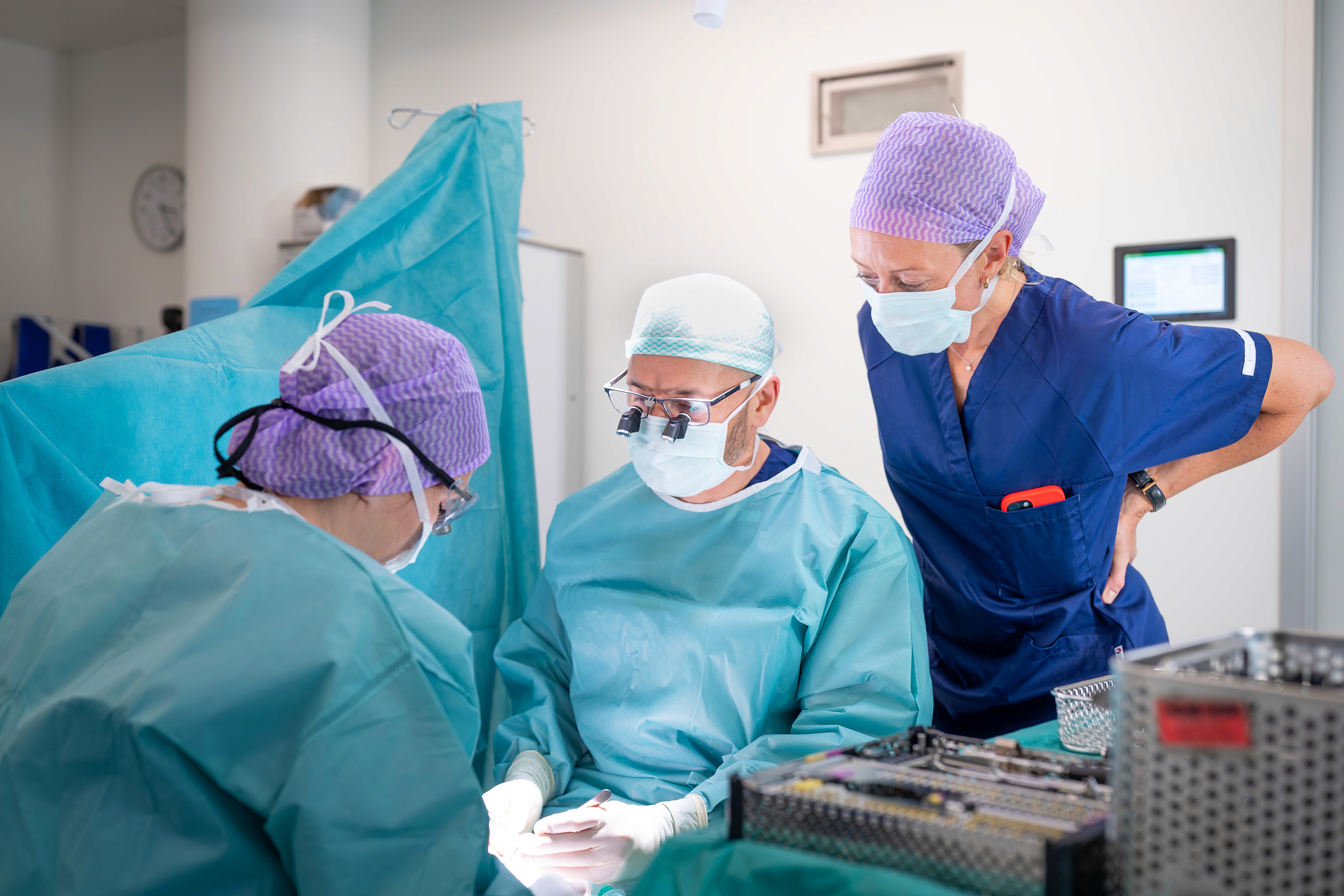 Drei medizinische Fachkräfte in OP-Kitteln und Masken führen in einem Operationssaal eine Operation durch, wobei eine steht und beobachtet, während zwei operieren. Im Vordergrund sind chirurgische Instrumente zu sehen.