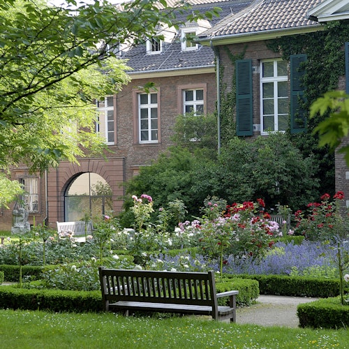 Ein ruhiger Garten mit Blumenbeeten, einer Holzbank und einem Backsteingebäude mit Fensterläden, die teilweise von Efeu bedeckt sind.