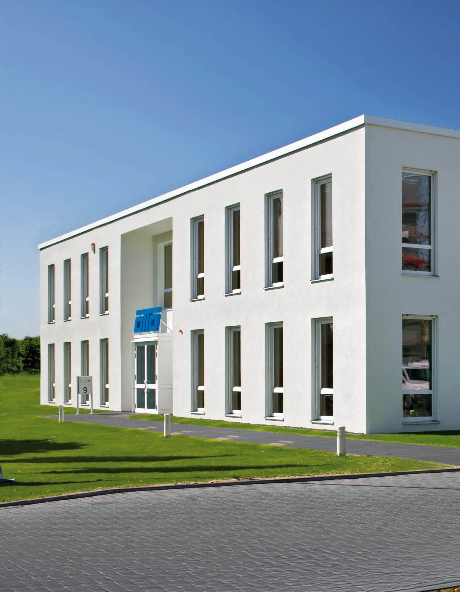 Modernes, weißes Bürogebäude mit vielen Fenstern und einem blauen Unternehmenslogo über dem Eingang.