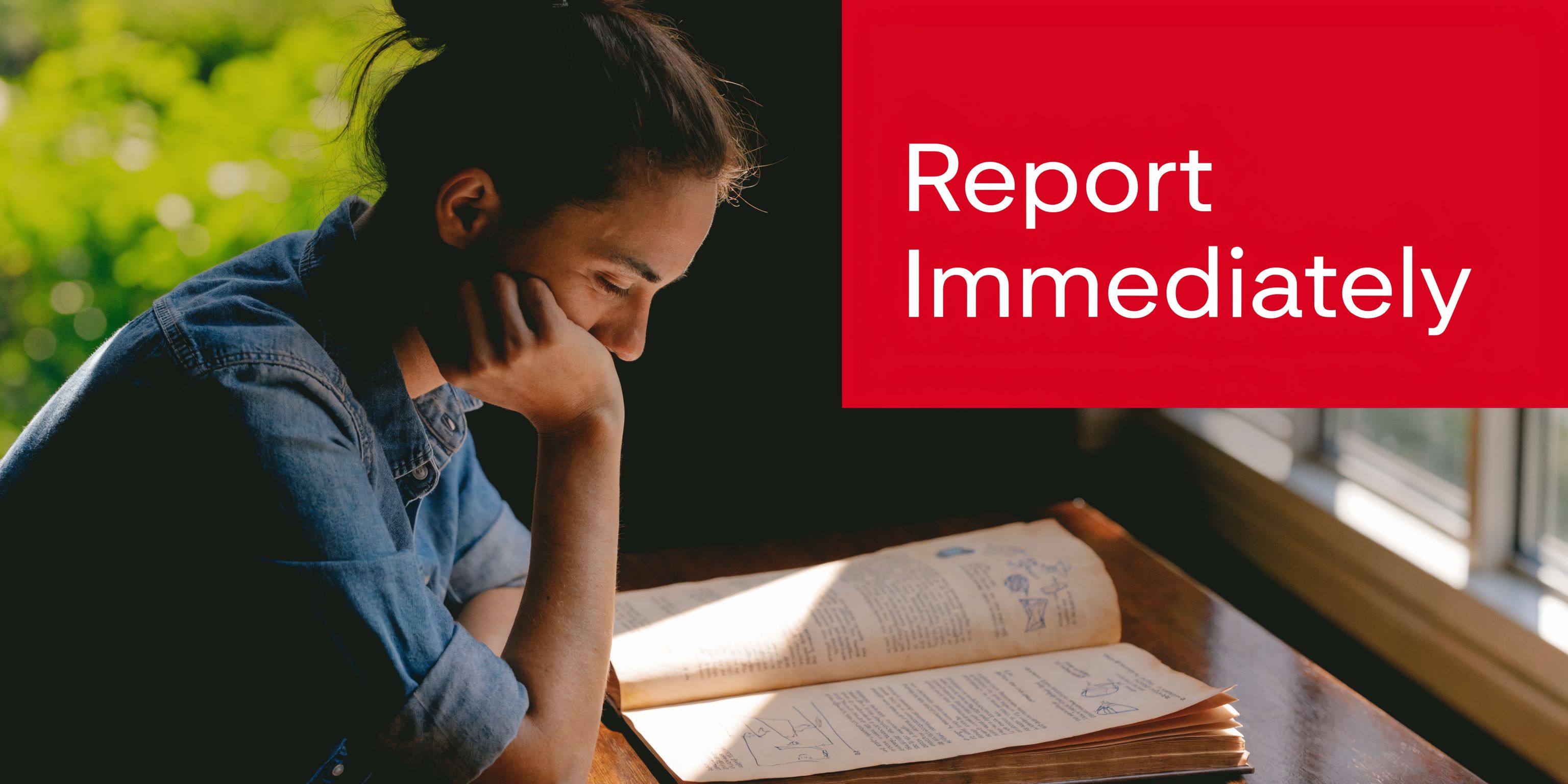 A person resting their chin on their hand while reading an open book at a wooden table.