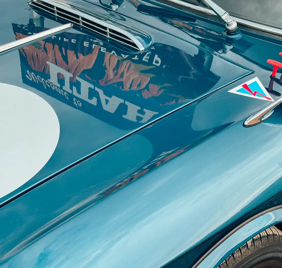 The hood of a blue Lancia Fulvia sports car, reflecting a “Welcome to Utah” sign.