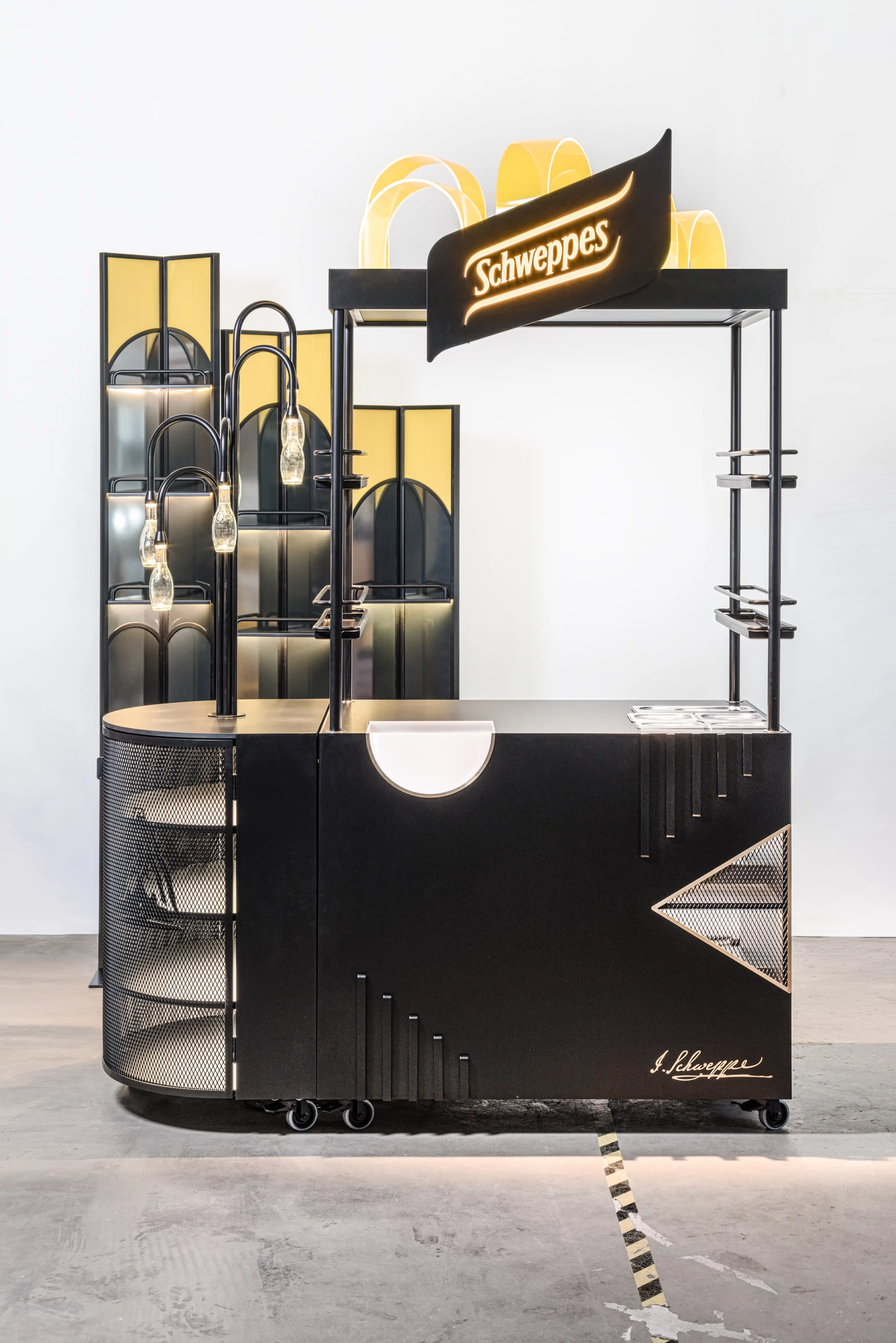 Mobile SCHWEPPES bartool in black with illuminated logo, golden Art Deco arches, bottle lamps and a folding screen with backlit shelves behind.