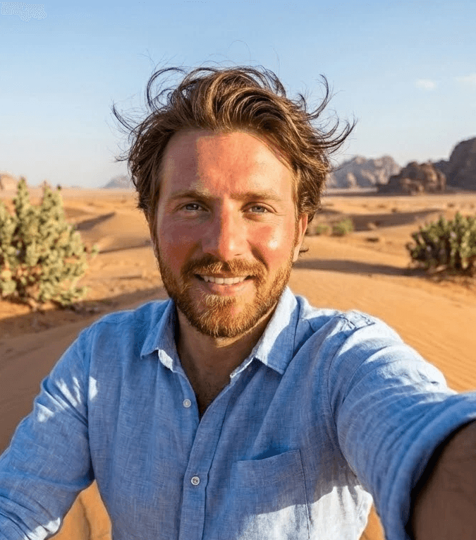 صورة سيلفي مُلتقطة من زاوية عالية لرجل مبتسم ذو شعر بني فاتح مُصفّف بالرياح ولحية مُهندمة، يقف في صحراء شاسعة. يرتدي قميصاً بلون أزرق فاتح مصنوع من الكتان ومزود بأزرار. تظهر في الخلفية كثبان رملية ذهبية متدحرجة، وشجيرات صحراوية خضراء نادرة، وجبال من الحجر الرملي الخشن تحت سماء زرقاء صافية وساطعة.