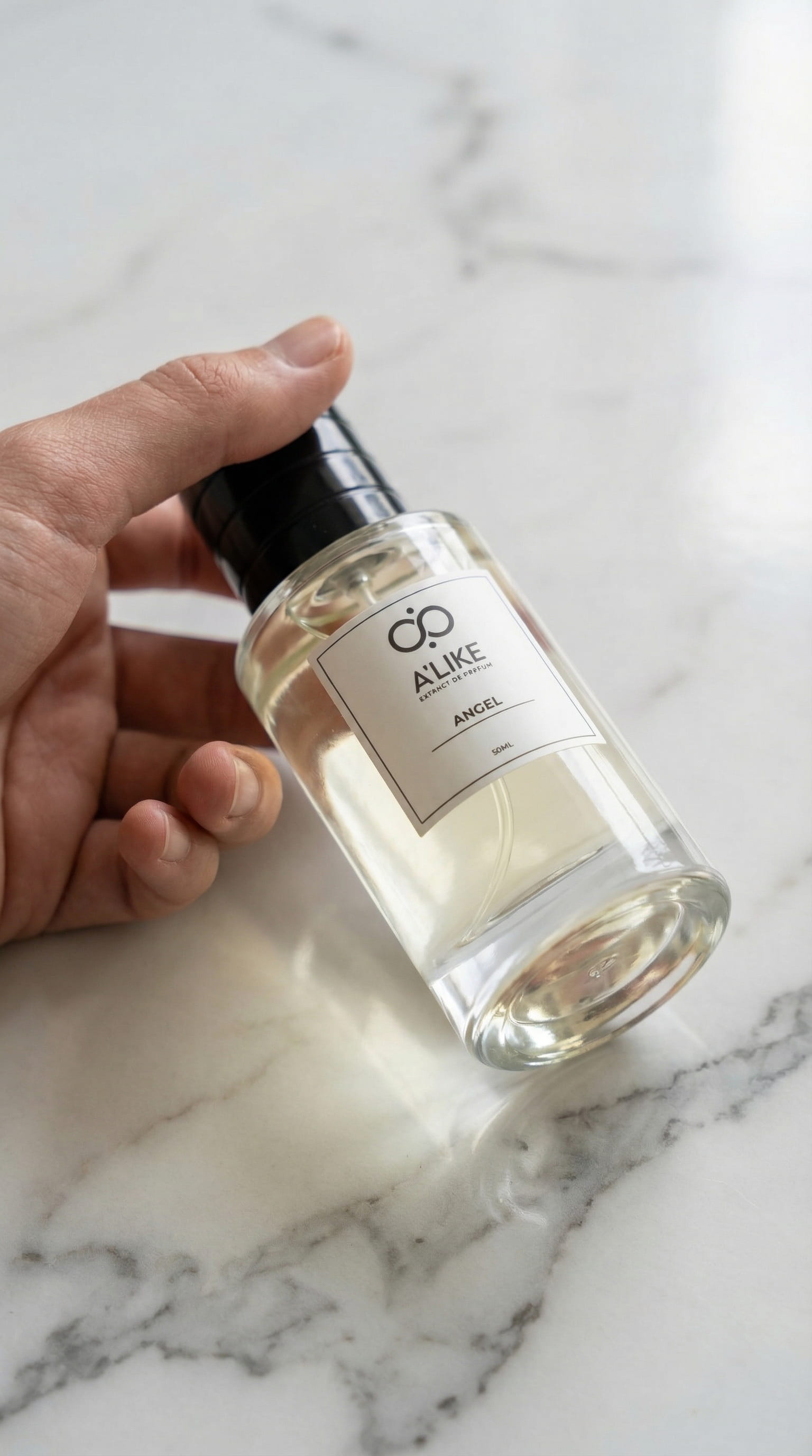 A hand holds a clear glass perfume bottle with a black cap. The bottle contains a light yellow liquid