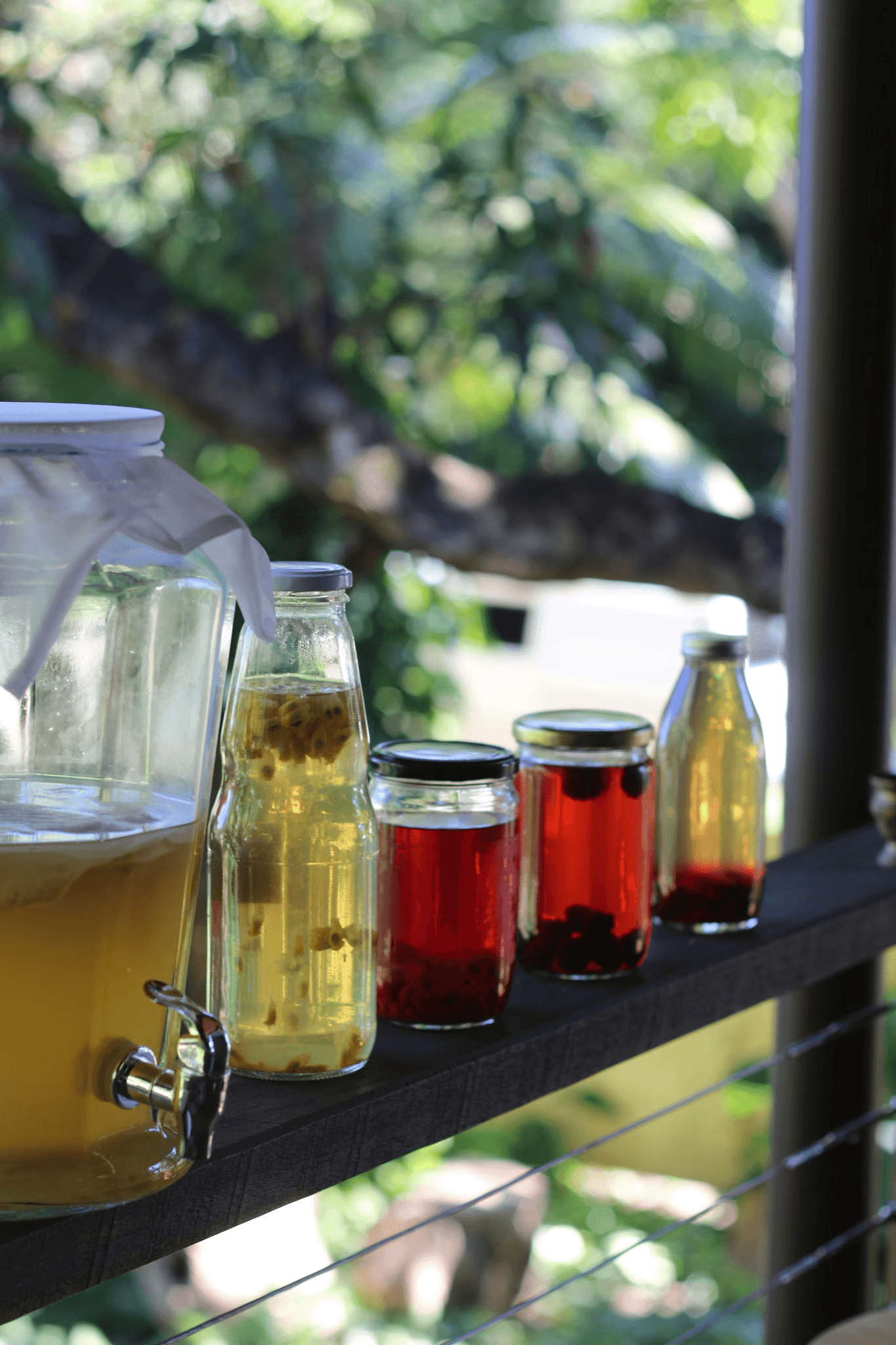 A selection of drinks in clear bottles, including light and dark liquids, placed on a wooden shelf outdoors.