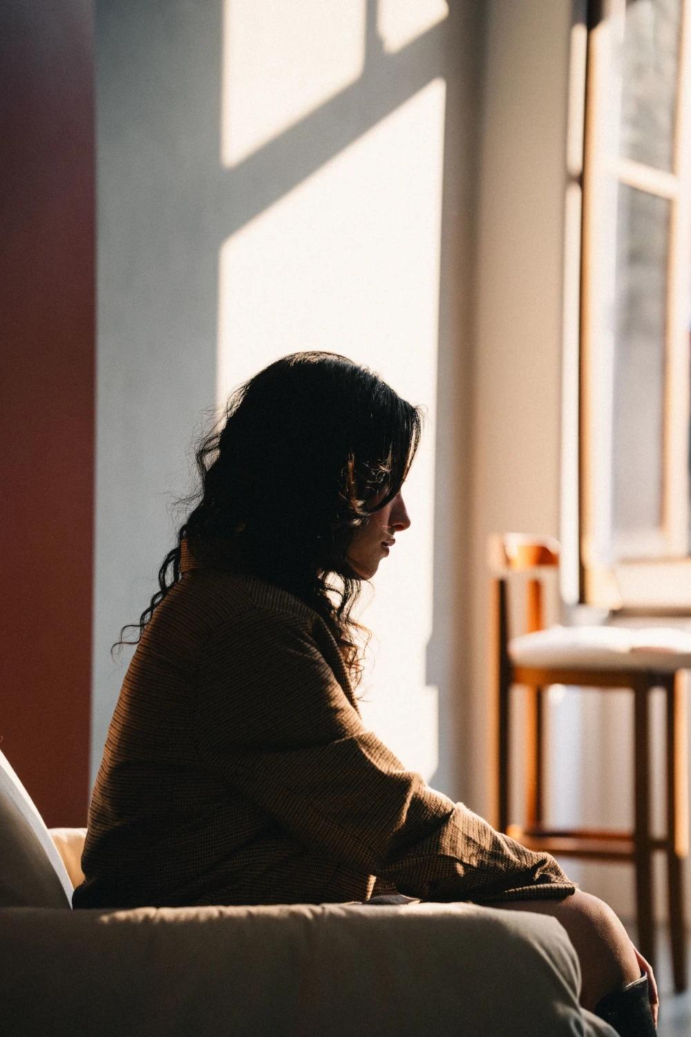 Woman sitting alone by a sunlit window, looking thoughtful and emotionally withdrawn.